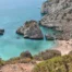 Praia de areia dourada rodeada de falésias calcárias, água azul-turquesa e rochas, com vegetação mediterrânica no topo.
