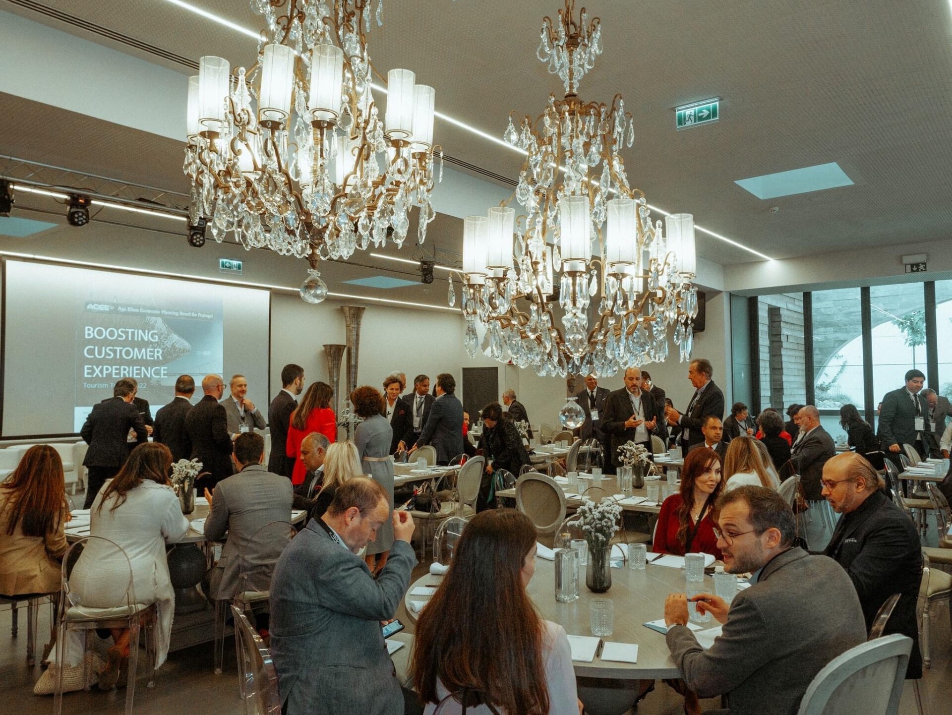 Conferência de negócios em sala elegante com candelabros, participantes em mesas e apresentação sobre experiência do cliente.
