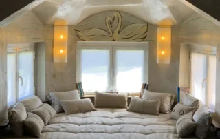 Quarto moderno com cama grande e almofadas bege, janelas amplas, iluminação quente e decoração minimalista.