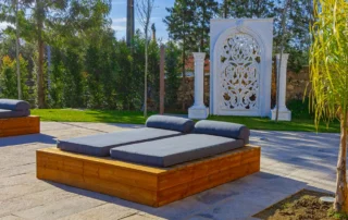 Espaço exterior com espreguiçadeiras de madeira e colchões cinzentos, decoração branca ornamentada e vegetação verde.