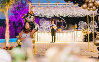 Mulher grávida em vestido branco com coroa segurando bolhas numa festa decorada com balões dourados e pretos à noite.