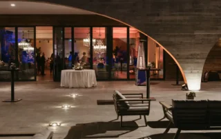 Evento nocturno num pátio com estrutura curva de madeira, mesas redondas, convidados e iluminação ambiente
