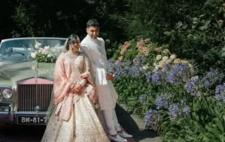 Casal de noivos junto a um carro clássico num jardim florido.